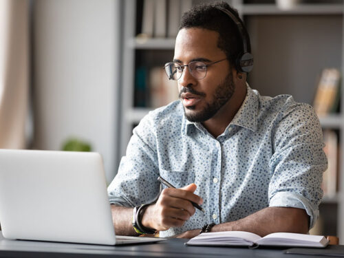 Focused young african businessman wear headphones study online watching webinar podcast on laptop listening learning education course conference calling make notes sit at work desk, elearning concept